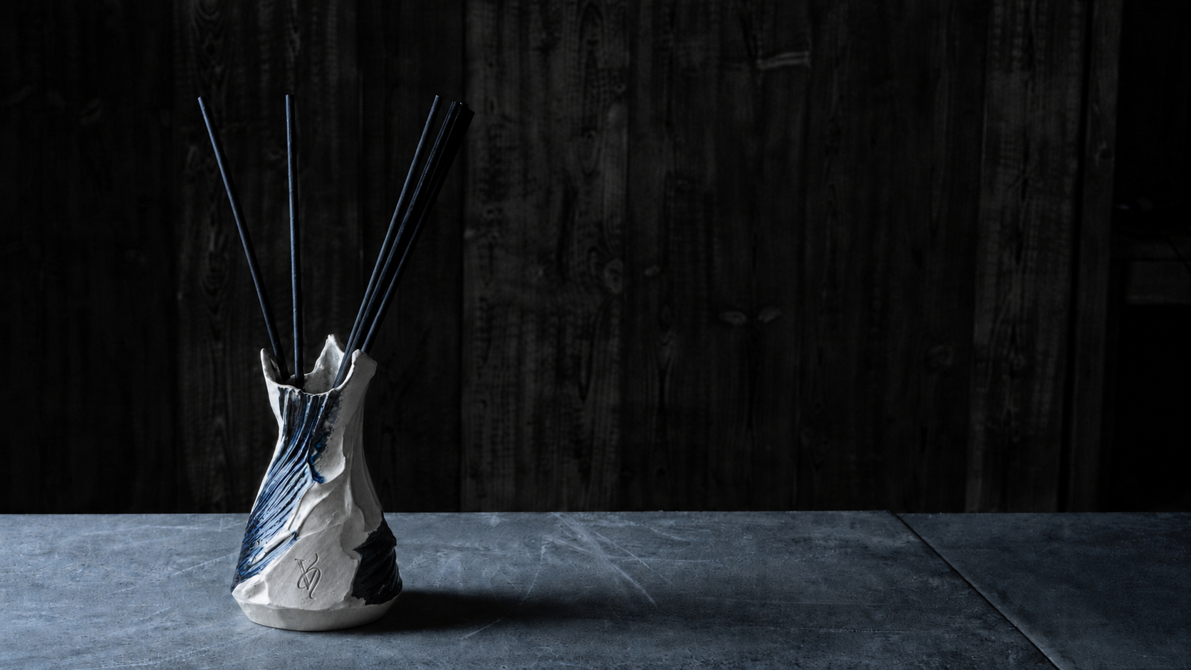 A hand-thrown Valdaire ceramic diffuser vessel with black reeds, on cool stone against a dark timber wall